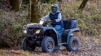 Driver on green Foreman going left on dirt road. Trees and bushes in background.