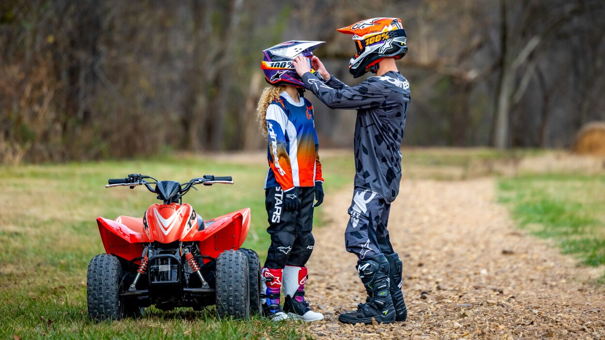 Young rider with Adult rider holding their helmet. Beside ATV.