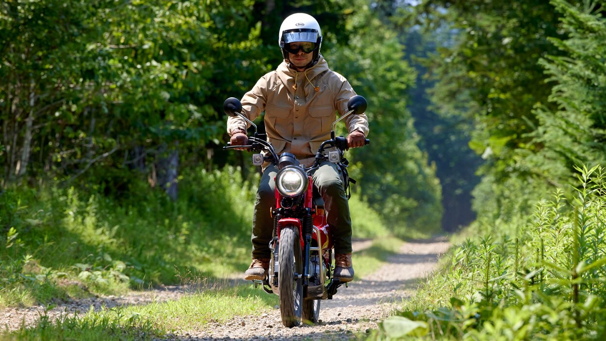 Rider with beige jacket on red bike riding through forest on stone road.