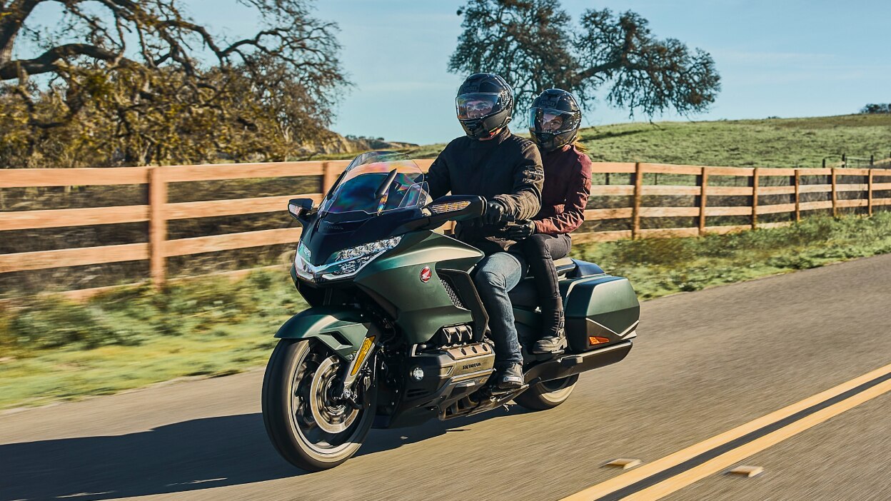 Two riders on green bike going left. Trees, field and fence in background.