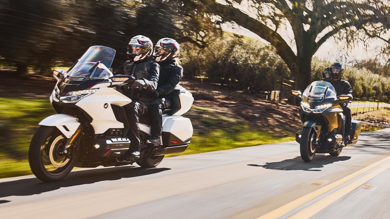 Two riders on white bike infront of two riders on green bike on road. Trees and bushes in background.