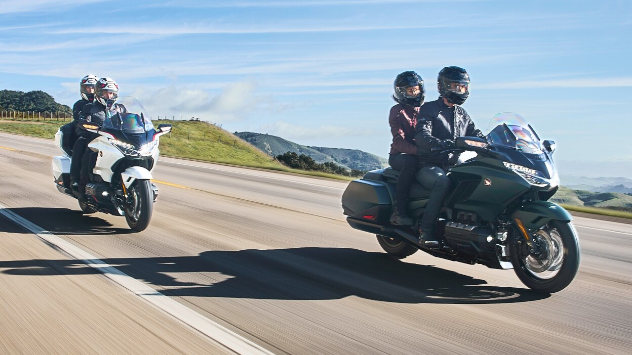 Two riders on green bike in front of two riders on white bike on road going right. Blue sky in background.