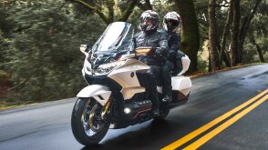 Two riders on white bike going left on road. Bushes and trees in background.