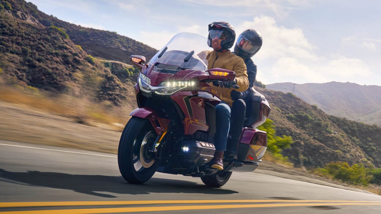 Rider and passenger on red Gold Wing Tour going left on road. Mountains in background.