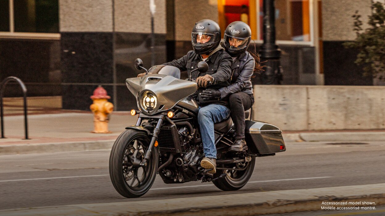 Rider and passenger on road in city landscape. Grey building in background. Accessorized model shown.