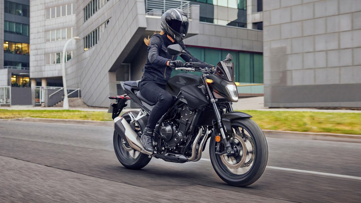 Rider with long hair driving on road. Grey building with large glass windows in background.