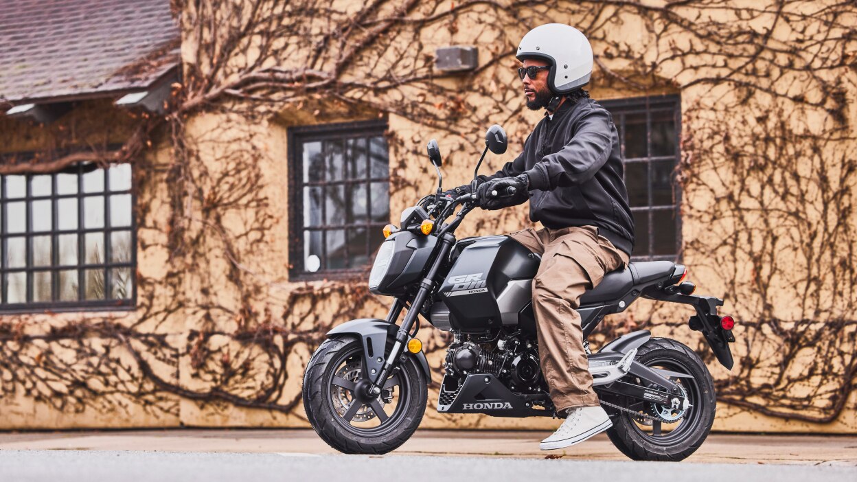 Grom with white helmet on handles stationary in front of building with vines on wall. Man at rear of bike.