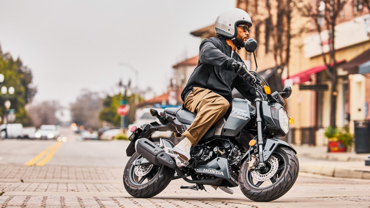 Rider on street with white helmet turning left on road on Grom. Buildings in background.