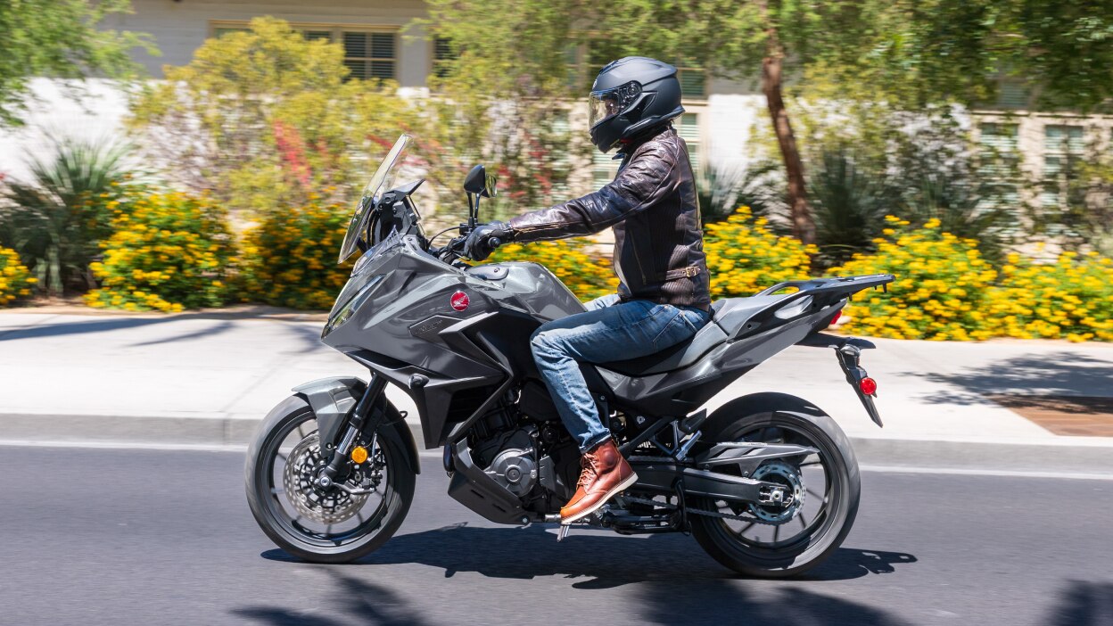 Rider on NT1100 going left on road. Pavement, colourful bushes and small trees in background.