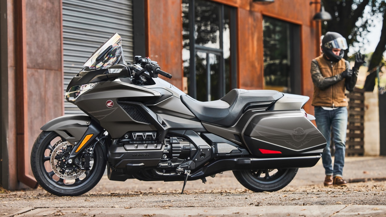 Left stationary image of Gold wing in front of red building. Person with Helmet on in background walking towards Gold Wing.