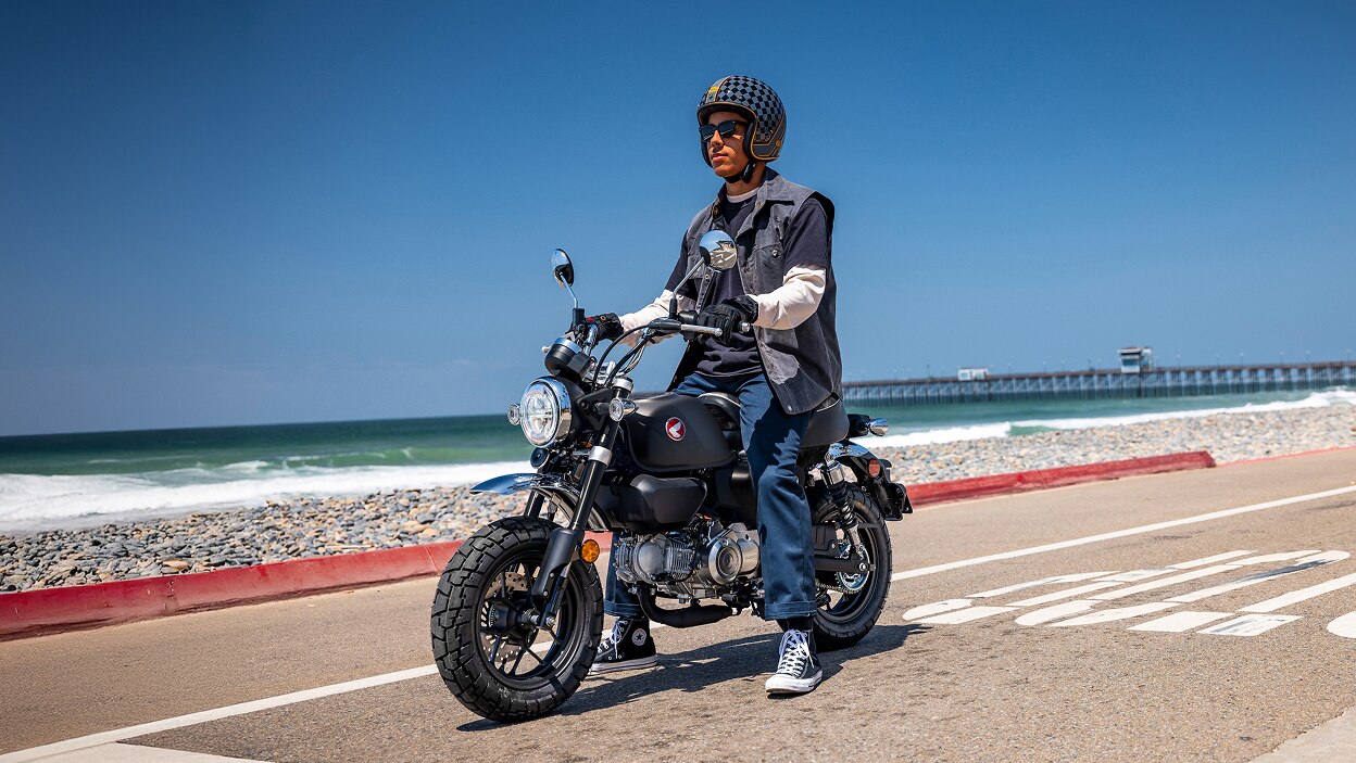 Rider on Motorbike. Stationary on road, facing left. Beach in background.