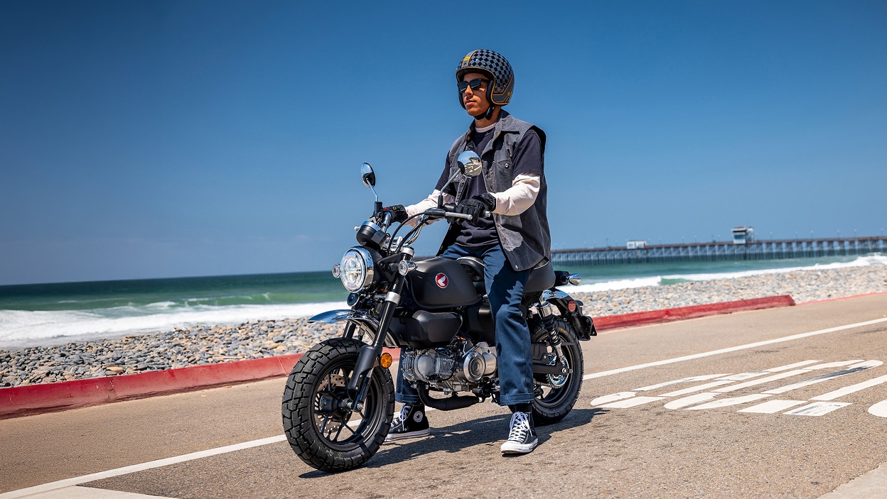 Rider on Motorbike. Stationary on road, facing left. Beach in background.