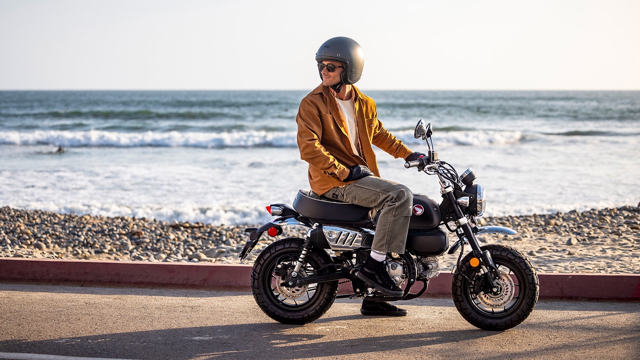 Rider on motorbike. Stationary on road, facing right. Beach in background.
