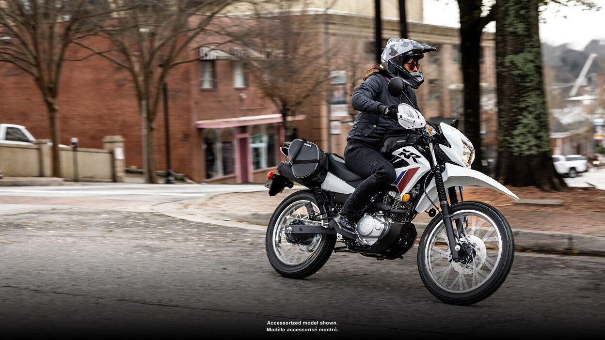 Rider stationary on CRF450L. City landscape and blue sky in background.