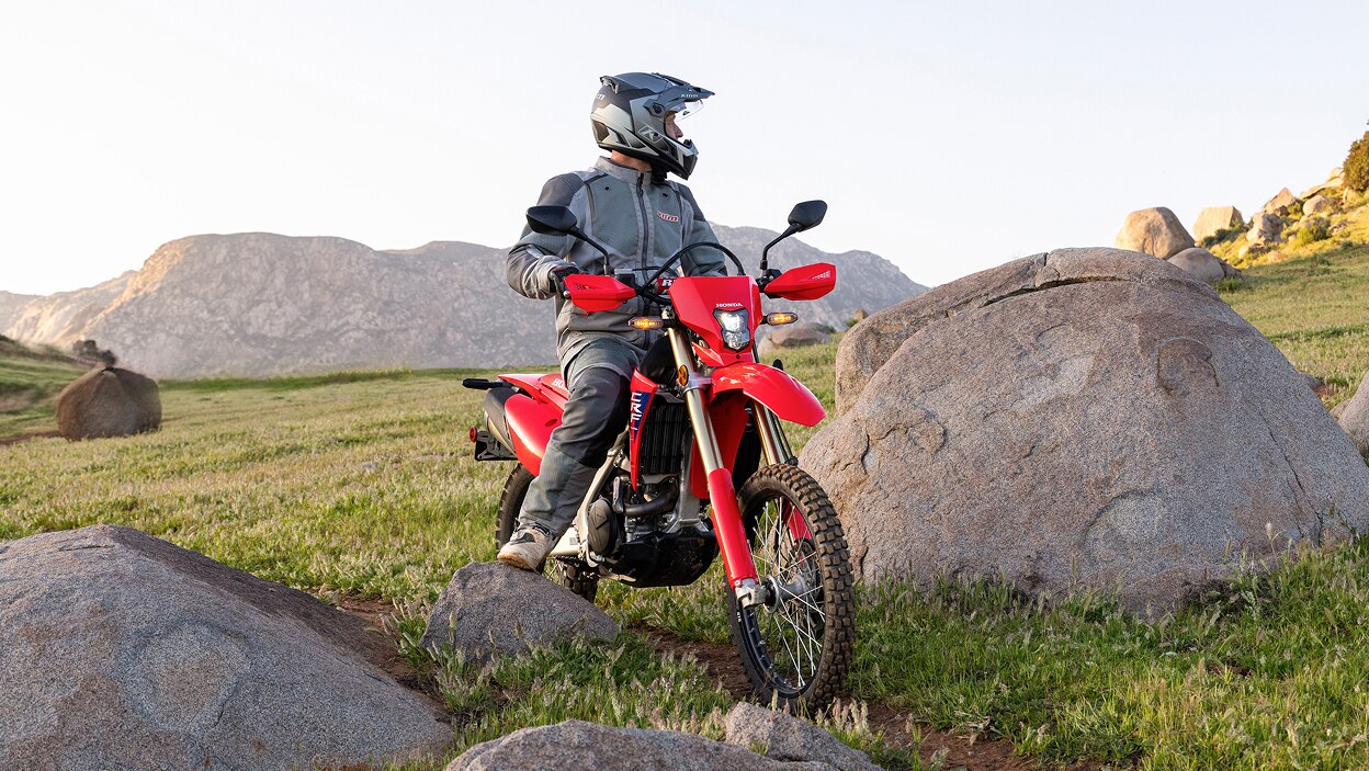 Rider standing on CRF450L. Rocky and green hills in background.