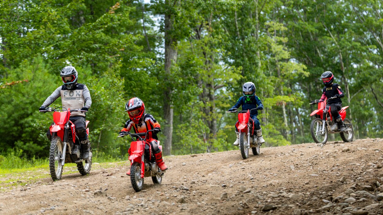 Four riders on red Honda trail bikes riding toward the camera on a sandy dirt track surrounded by dense green trees.