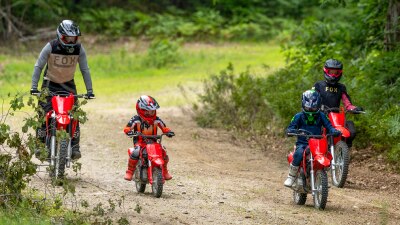 Two young riders on small red dirt bikes navigating a dirt trail through the woods.