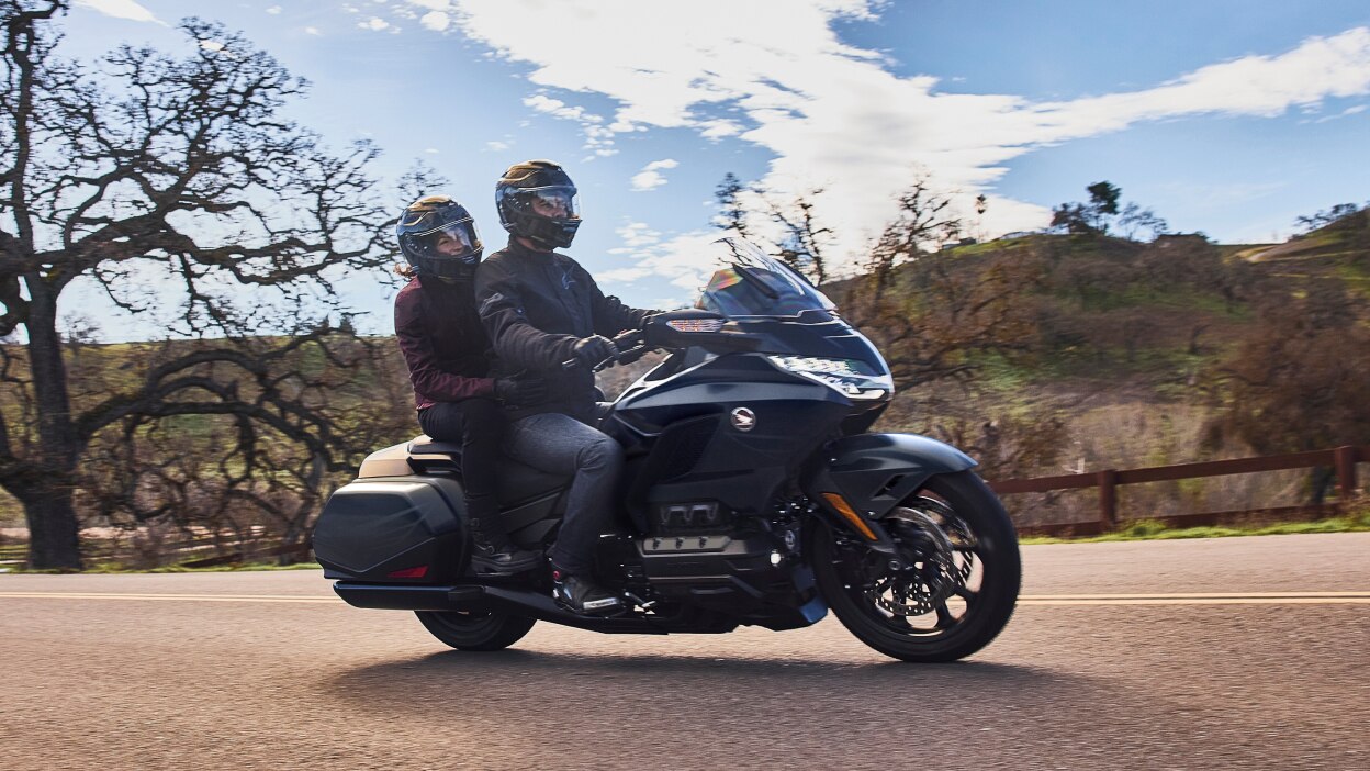 Two people riding a black touring motorcycle on a road near bare trees and hills.