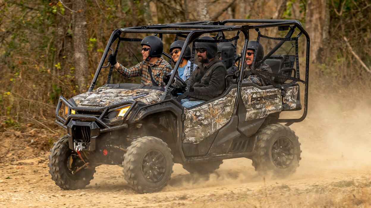 Four people in camo sxs going left on dirt track. Forrest background.